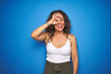 Kıvırcık saçlı orta yaş yaşlı kadın mavi izole arka plan üzerinde duran el gülümseyerek ok jest yapıyor, mutlu yüz ile parmaklardan bakan göz.