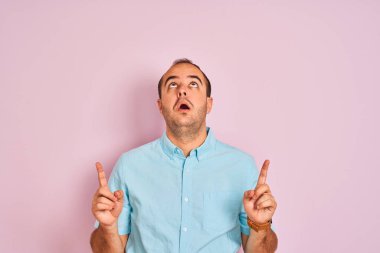 Izole pembe arka plan üzerinde duran mavi rahat gömlek giyen genç adam şaşırttı ve yukarı bakarak ve parmakları ile işaret ve kolları kaldırdı şaşırttı.
