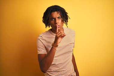 Çizgili t-shirt giyen dreadlocks ile Afro adam dudaklarında parmak ile sessiz olmak isteyen izole sarı arka plan üzerinde duruyor. Sessizlik ve gizli kavram.