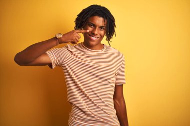 Çizgili t-shirt giyen dreadlocks ile Afro adam izole sarı arka plan üzerinde duran yüz ve burun el parmağı ile işaret, neşeli gülümseyen. Güzellik konsepti
