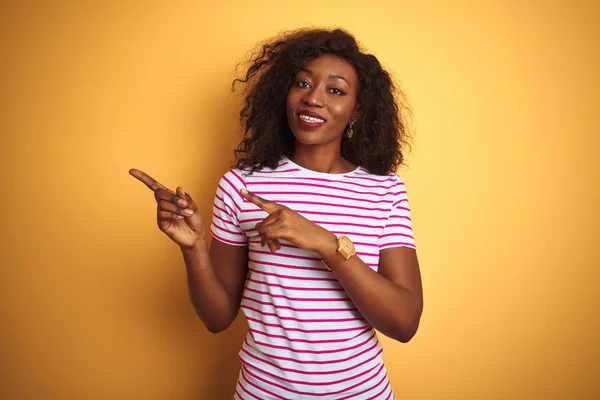Young african american woman wearing striped t-shirt over isolated yellow background smiling and looking at the camera pointing with two hands and fingers to the side.