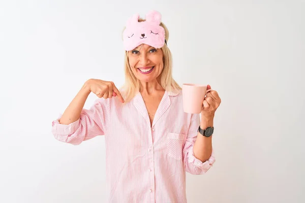 Middle age woman wearing sleep mask pajama drinking coffee over isolated white background with surprise face pointing finger to himself