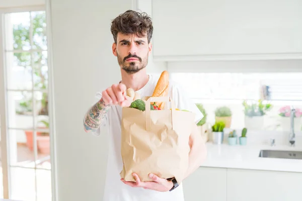 Young man holding paper bag of fresh groceries from the supermarket pointing with finger to the camera and to you, hand sign, positive and confident gesture from the front