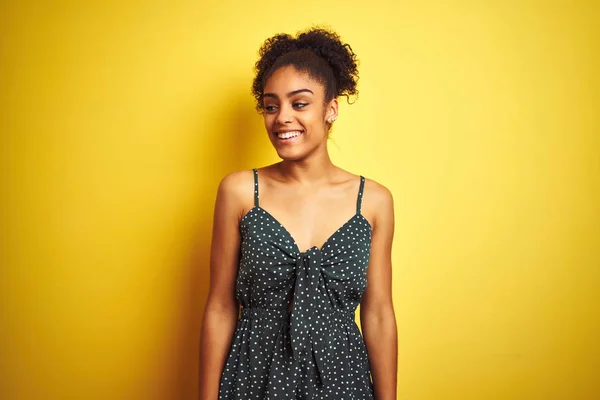 African american woman wearing summer casual green dress over isolated yellow background looking away to side with smile on face, natural expression. Laughing confident.