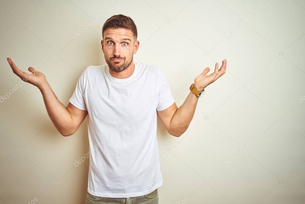 Young handsome man wearing casual white t-shirt over isolated background clueless and confused expression with arms and hands raised. Doubt concept.