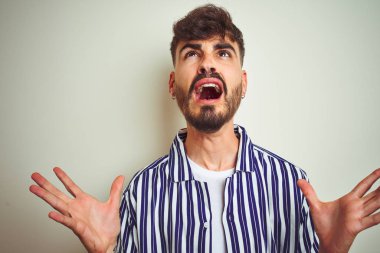 Dövmeli genç adam çizgili gömlek giyen izole beyaz arka plan üzerinde duran deli ve deli bağırarak ve agresif ifade ve kollar kaldırdı ile bağırıyor. Hayal kırıklığı kavramı.