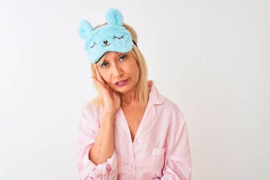 Orta yaş kadın mavi uyku maskesi ve pijama giyen izole beyaz arka plan üzerinde yorgun görünümlü ve çapraz kollar ile depresyon sorunları ile sıkılmış düşünme.