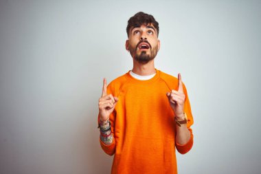 Izole beyaz arka plan üzerinde duran turuncu kazak giyen dövme giyen genç adam şaşırttı ve yukarı bakarak ve parmakları ile işaret ve kolları kaldırdı şaşırttı.