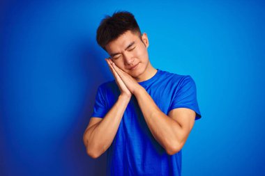 İzole mavi arka plan üzerinde duran t-shirt giyen genç Asyalı çinli adam yorgun rüya ve kapalı gözlerle gülümseyerek birlikte elleri ile poz.