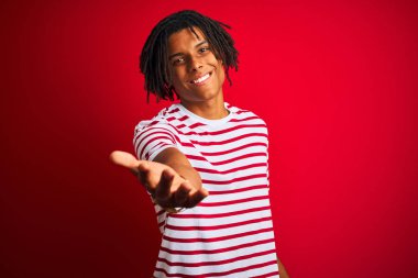 İzole kırmızı arka plan üzerinde duran çizgili t-shirt giyen dreadlocks giyen genç afro adam yardım ve kabul veren palmiye el sunan neşeli gülümseyerek.