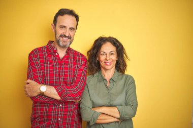 İzole sarı arka plan üzerinde güzel orta yaş çift kamera bakarak çapraz silah ile gülümseyen mutlu yüz. Pozitif kişi.