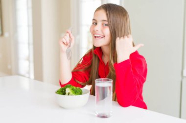 Güzel genç kız çocuk taze brokoli yeme ve içme suyu işaret ve mutlu yüzü gülümseyerek yan başparmak ile gösteren