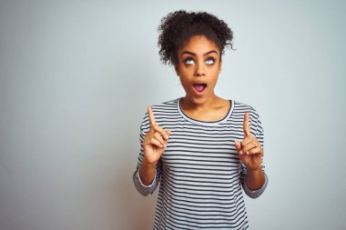 Izole beyaz arka plan üzerinde duran lacivert çizgili t-shirt giyen Afro-Amerikan kadın şaşırttı ve yukarı bakarak ve parmakları ile işaret ve kolları kaldırdı şaşırttı.