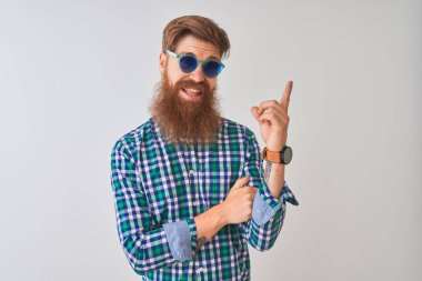 Genç kızıl İrlandalı adam yüzünde büyük bir gülümseme ile izole beyaz arka plan üzerinde rahat gömlek ve güneş gözlüğü giyen, kameraya bakarak yan el ve parmak ile işaret.