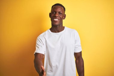 Beyaz tişört giyen genç afro-amerikan adam izole sarı arka plan üzerinde ayakta selamlama ve karşılama olarak dostça sunan el sıkışma gülümseyerek. Başarılı iş.