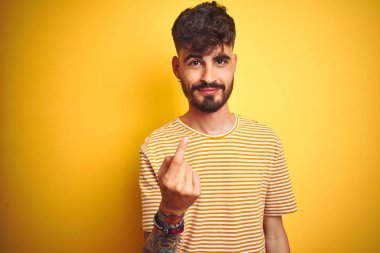 Çizgili t-shirt giyen genç adam izole sarı arka plan Beckoning üzerinde duran el davet eden mutlu ve gülümseyerek buraya jest gel