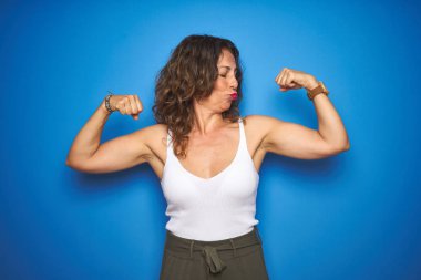 Kıvırcık saçlı orta yaş yaşlı kadın mavi izole arka plan üzerinde duran kol kasları gururla gülümseyen gösteren. Fitness konsepti.