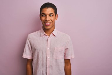 Genç yakışıklı arap adam izole pembe arka plan üzerinde duran rahat gömlek giyen yan bakarak gülümseyerek ve düşünme uzak bakıyor.