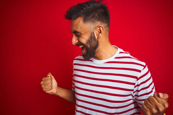 年轻的印度男子穿着条纹T恤站在孤立的红色背景非常高兴和兴奋做胜利者手势与手臂抬起 微笑和尖叫的成功 庆典概念 — 图库照片