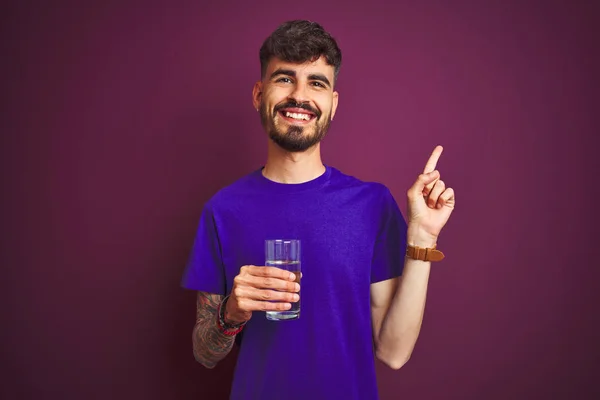 Молодий Чоловік Татуюванням Склянку Води Стоїть Над Ізольованим Фіолетовим Тлом — стокове фото