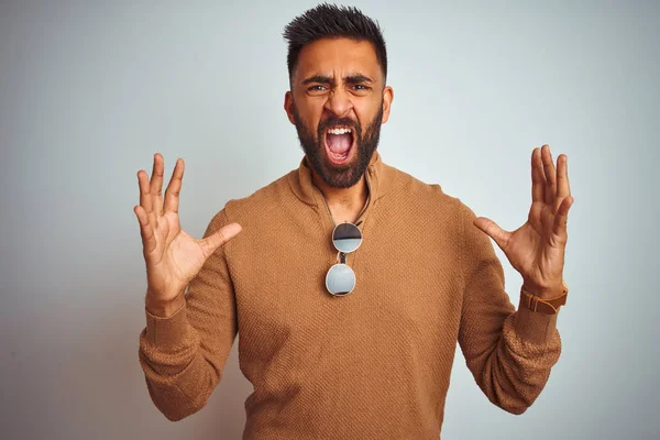 Indian man wearing elegant sweater and sunglasses standing over isolated white background crazy and mad shouting and yelling with aggressive expression and arms raised. Frustration concept.