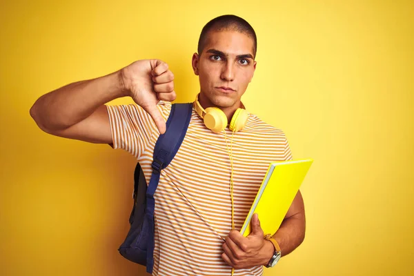 怒った顔で黄色の孤立した背景の上にヘッドフォンとバックパックを身に着けている若い学生の男 親指を下に嫌いを示す否定的なサイン 拒絶概念 — ストック写真