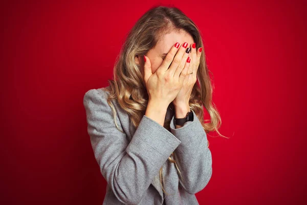 Young beautiful business woman wearing elegant jacket standing over red isolated background with sad expression covering face with hands while crying. Depression concept.