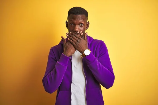 African american man wearing purple sweatshirt standing over isolated yellow background shocked covering mouth with hands for mistake. Secret concept.