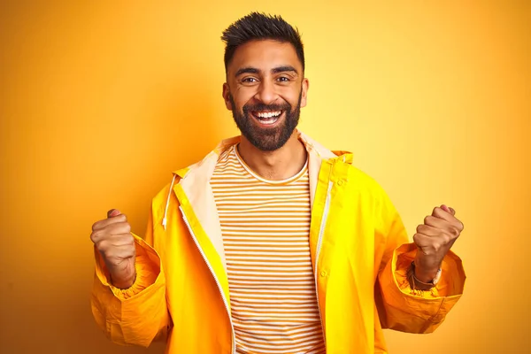 Young indian man wearing raincoat standing over isolated yellow background celebrating surprised and amazed for success with arms raised and open eyes. Winner concept.