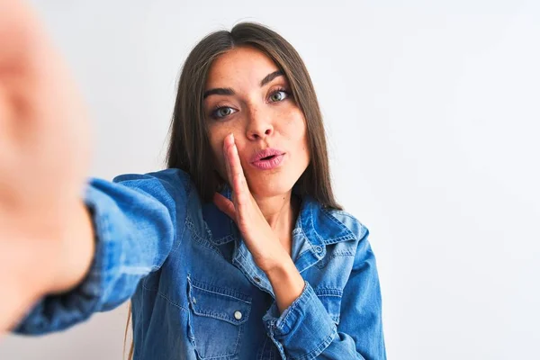 Beautiful woman wearing denim shirt make selfie by camera over isolated white background hand on mouth telling secret rumor, whispering malicious talk conversation