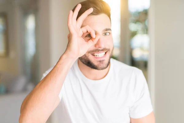 年轻英俊的男人穿着休闲白色 T恤在家里做确定的手势与手微笑 眼睛通过手指看着快乐的脸 — 图库照片