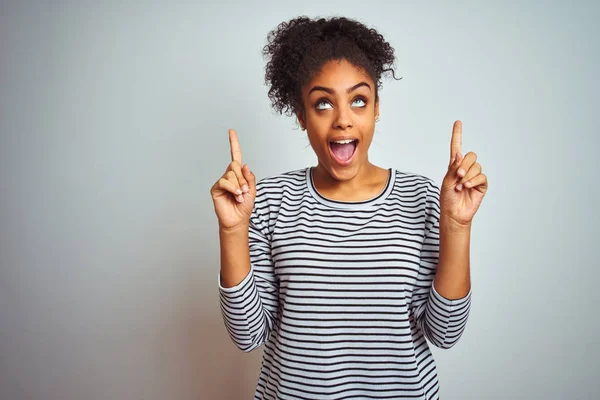 African american woman wearing navy striped t-shirt standing over isolated white background smiling amazed and surprised and pointing up with fingers and raised arms.