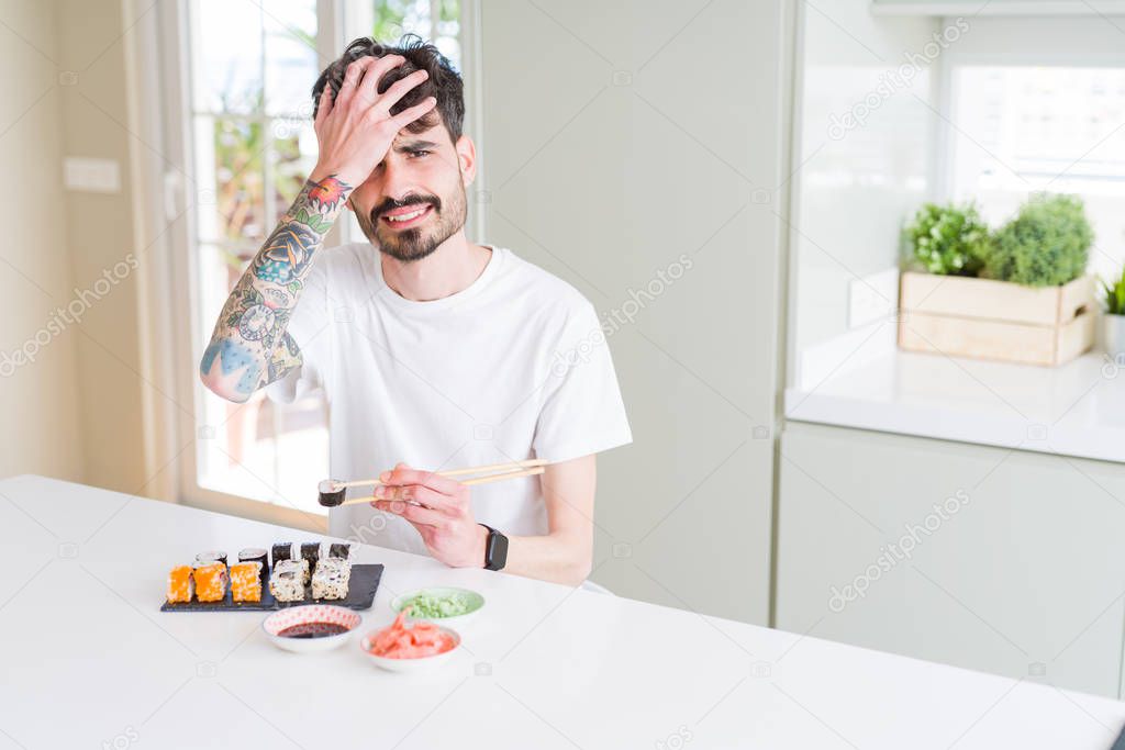 Young man eating asian sushi from home delivery stressed with hand on head, shocked with shame and surprise face, angry and frustrated. Fear and upset for mistake.