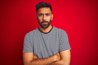 Genç yakışıklı hintli adam izole kırmızı arka plan şüpheci ve sinir üzerinde lacivert çizgili t-shirt giyen, çapraz kolları ile yüzünde onaylamayan ifade. Negatif kişi.