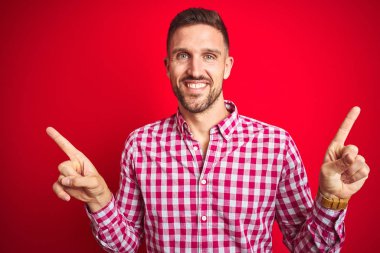 Kırmızı izole arka plan üzerinde genç yakışıklı adam farklı yönlere parmakları ile işaret emin gülümseyerek. Reklam için kopya alanı
