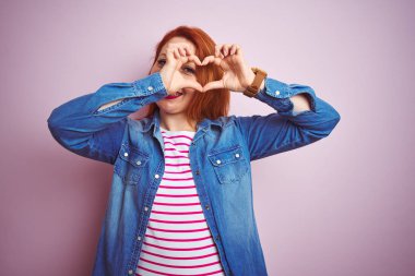 Denim gömlek giyen güzel kızıl saçlı kadın ve izole pembe arka plan üzerinde çizgili t-shirt el ve parmaklar işareti bakarak gülümseyerek kalp şekli yapıyor