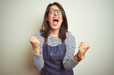 Genç güzel iş kadını izole arka plan üzerinde mağaza üniforma önlük giyen çok mutlu ve heyecanlı kolları kaldırdı ile kazanan jest yapıyor, gülümseyerek ve başarı için çığlık. Kutlama konsepti.