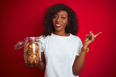 Genç afrikalı amerikalı kadın izole kırmızı arka plan üzerinde kurabiye kavanozu tutan çok mutlu yan el ve parmak ile işaret