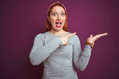 Güzel kızıl saçlı kadın gri t-shirt giyen ve izole mor arka plan üzerinde diadem şaşırttı ve el ile sunarken kameraya gülümseyerek ve parmak ile işaret.