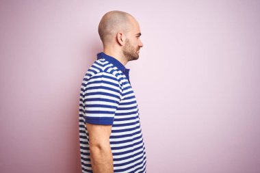 Sakallı genç kel adam pembe izole arka plan üzerinde rahat çizgili mavi t-shirt giyen yan bakarak, kendine güvenen gülümseme ile doğal yüz ile profil poz rahat.