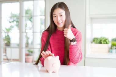Güzel Asyalı kadın ok işareti yapıyor büyük bir gülümseme ile mutlu piggy çanta içinde bir sikke koyarak, parmakları ile başparmak, mükemmel işareti