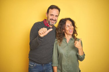 Izole sarı arka plan üzerinde güzel orta yaş çift mutlu yüz görünümlü ve başparmak ile yan işaret gülümseyerek.