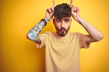 Çizgili t-shirt giyen dövme giyen genç adam boğa boynuzları gibi baş üzerinde parmak ile komik jest yapıyor izole sarı arka plan üzerinde duran