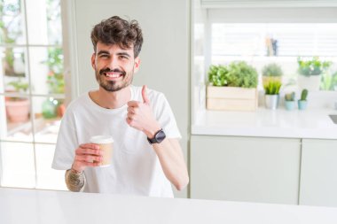 Genç adam sabah bir kağıt fincan bir kahve içme ok işareti yapıyor büyük bir gülümseme ile mutlu, parmakları ile başparmak, mükemmel işareti