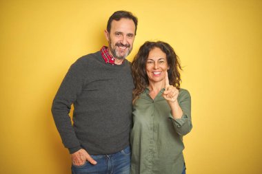 Izole sarı arka plan üzerinde Güzel orta yaş çift gösteren ve emin ve mutlu gülümseyerek parmak sayısı bir ile işaret.