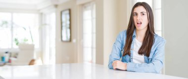 Evde beyaz masada oturan güzel genç kadının geniş açılı resmi Şok yüzünde, şüpheci ve alaycı görünümlü, açık ağız ile şaşırttı