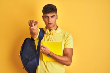 Kamera ve size parmak ile işaret izole sarı arka plan üzerinde sırt çantası kulaklık dizüstü bilgisayar giyen Hintli öğrenci adam, el işareti, önden olumlu ve kendinden emin jest