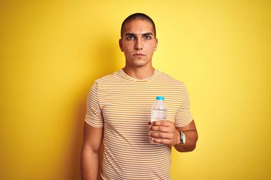 Genç yakışıklı adam ciddi düşünme akıllı yüz üzerinde emin bir ifade ile sarı izole arka plan üzerinde plastik su şişesi tutan