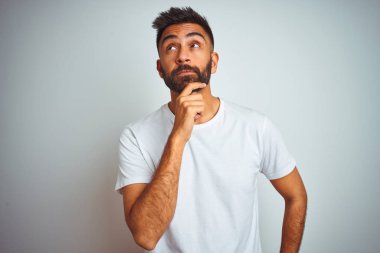 İzole beyaz arka plan üzerinde duran tişört giyen genç hintli adam çenesi üzerinde soru, dalgın ifade yi düşünüyor. Düşünceli bir yüzle gülümsüyor. Şüphe kavramı.