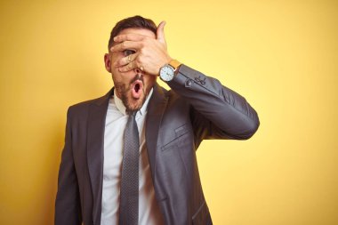 Sarı izole arka plan üzerinde genç yakışıklı iş adamı şok yüz ve gözleri el ile kapsayan gözetleme, utanmış ifade ile parmaklarından bakarak.